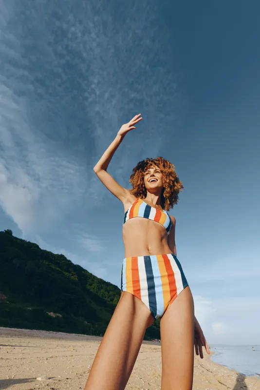 Woman on beach, striped swimsuit and curly hair, arms raised to the summer sky, warm sunlight, relaxed posture, ocean horizon, nature colors, tranquil mood with soft sea breeze today