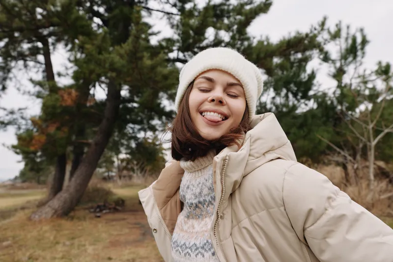 Winter scene with a smiling woman outdoors in a warm jacket and beanie, enjoying the park breeze and fresh air with a cozy scarf and relaxed mood.
