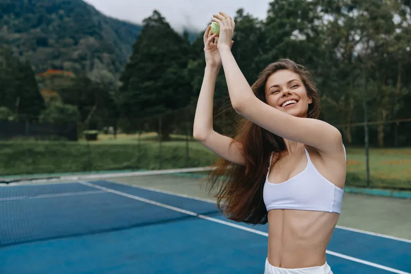 Young athletic woman playing tennis outdoors, smiling joyfully with a tennis ball, wearing a stylish white sports outfit on a green court