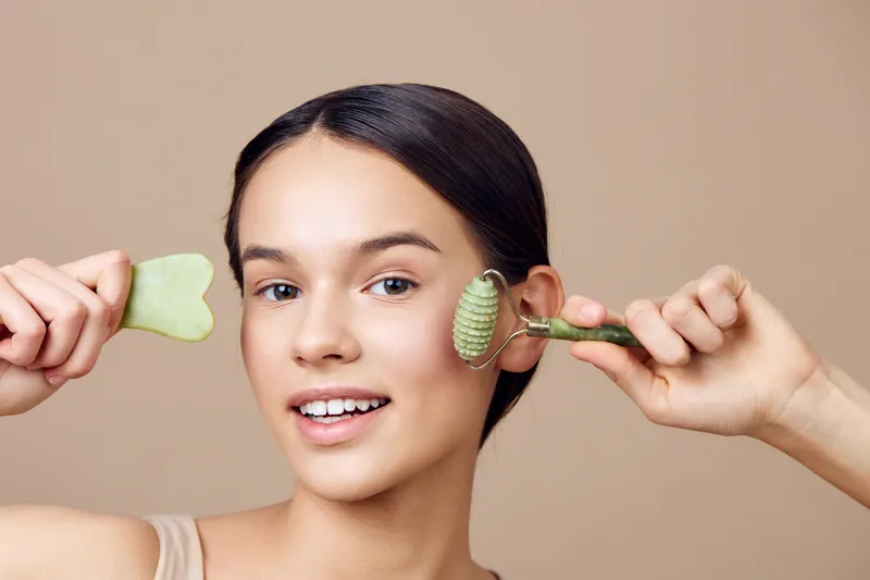 Young woman using facial rollers for skincare in a neutral background, showcasing healthy beauty rituals and self care concepts