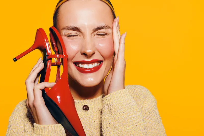 Happy woman with red lipstick holds red high heel shoe near face, eyes closed, smiling. Bright yellow background and cozy sweater create cheerful and vibrant fashion concept.