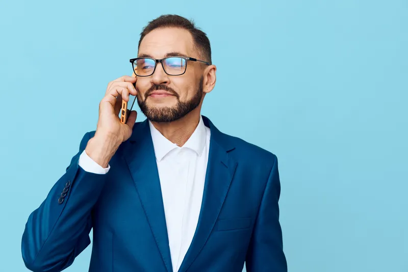 A confident business professional in a blue suit talks on a smartphone, wearing glasses, with a subtle smile, set against a solid blue background for an isolated color scene.