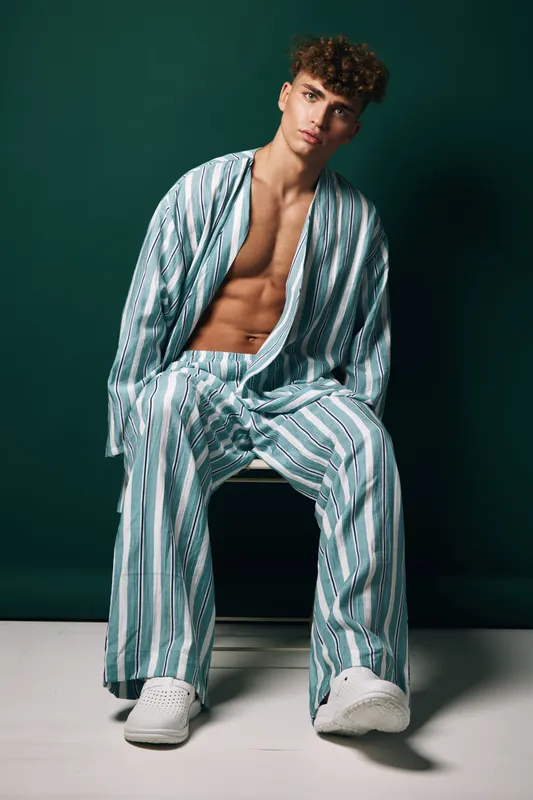Young man with curly hair wearing striped loose pajamas and white sneakers sitting on chair against dark green background in studio. Casual fashion, relaxed pose and modern style.