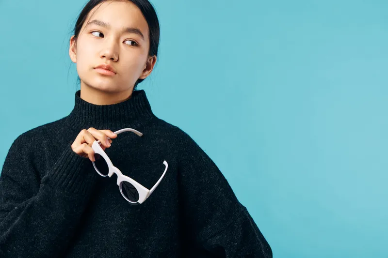 Young woman in black sweater holds white sunglasses with thoughtful expression on blue background. Portrait of casual lifestyle, fashion accessory and modern style with confident look.