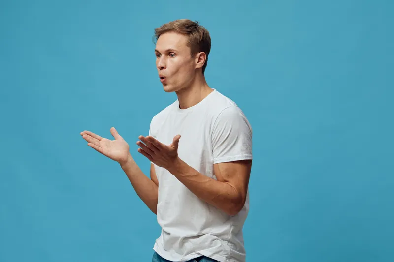 Confused young man with short hair wearing casual white t-shirt expression of surprise or questioning isolated on solid blue background studio portrait. People lifestyle concept