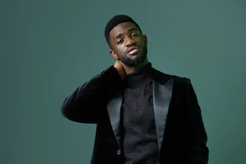 Уверенный мужчина в чёрном костюме и водолазке стоит в эффектной позе на фоне ярко-зелёного фона, излучая профессионализм и стиль
