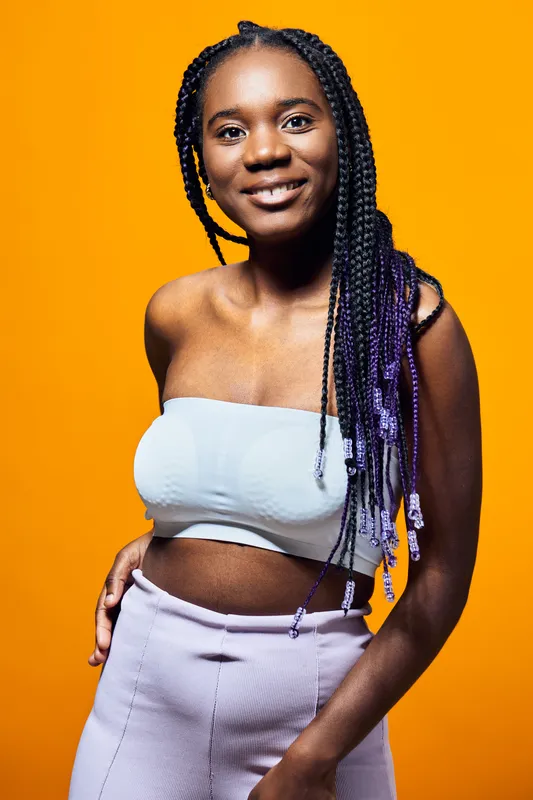 Confident young black woman with braided hair in a stylish outfit poses against a vibrant orange background, showcasing fashion and self expression