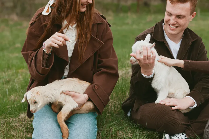 Casual couple outdoors with a playful goat kid, relaxed moment on sunlit grass, gentle interaction, candid lifestyle scene, warm tones, natural setting, wholesome bonding and genuine connection
