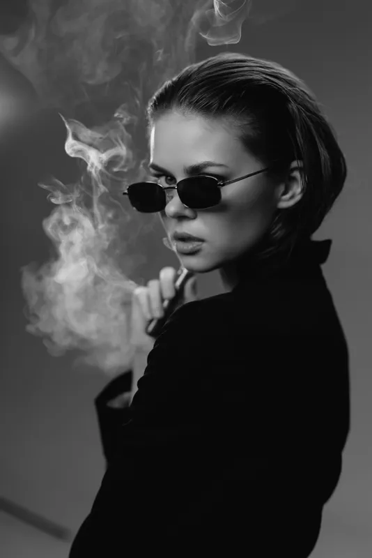 Black and white portrait of a stylish woman wearing sunglasses with smoke around her, creating a mysterious and edgy atmosphere.