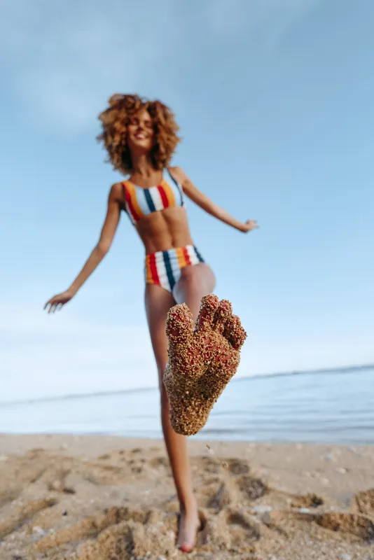 Woman with curly hair in striped bikini running on sandy beach, blue ocean waves behind, summer vacation mood, playful stride, heart shaped sand decoration in foreground, outdoors