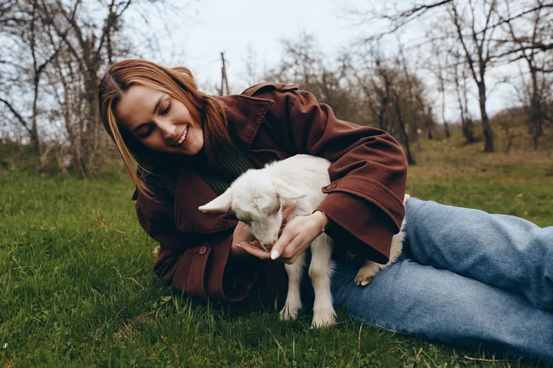Cheerful woman outdoors, lying on grass with a gentle goat, kid nearby, warm smile, relaxed pose, soft natural light, casual jacket, autumnal park setting, close bond, nature atmosphere