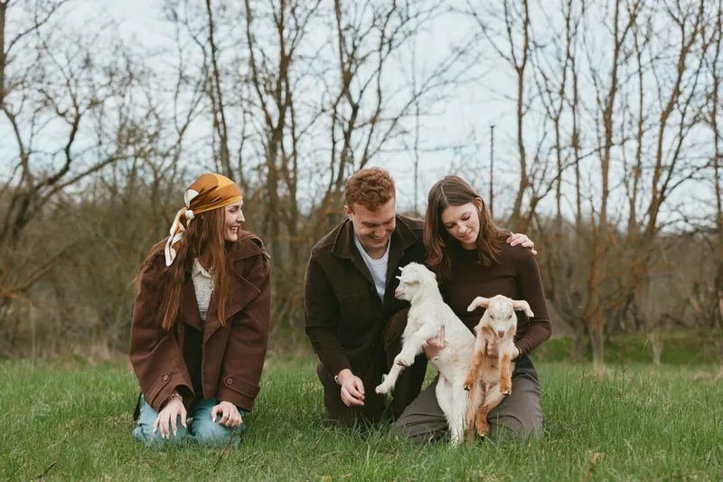 Young man and two women enjoy a spring day outdoors while playing with two baby goats in a grassy field surrounded by bare trees under an overcast sky