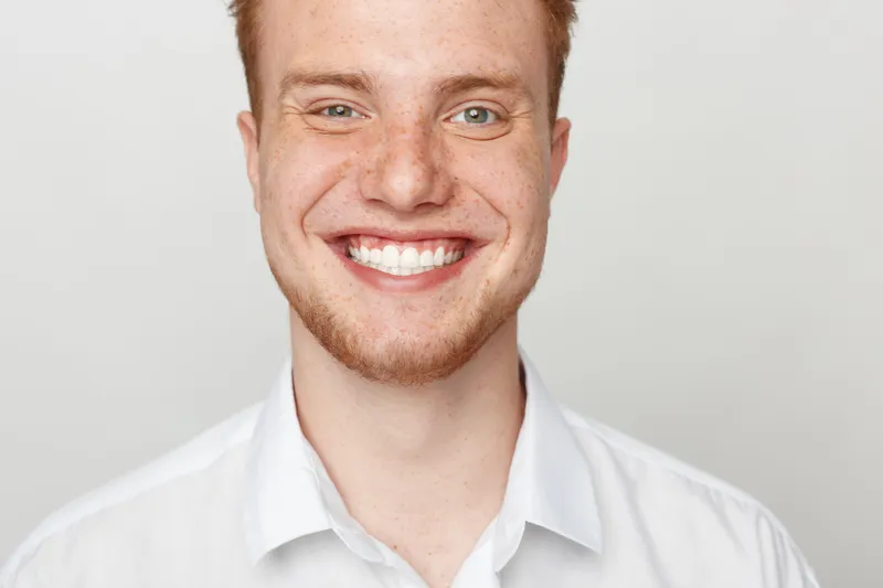 Smiling young man with red hair and beard wearing white shirt looks at camera. Close-up portrait on light background. Concept of happiness, confidence, and positive emotions.