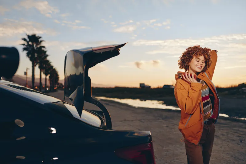 Curly haired woman in orange jacket standing beside car near beach road at sunset, lifestyle travel concept, warm golden light, casual outdoor portrait, evening breeze, soft mood, serene