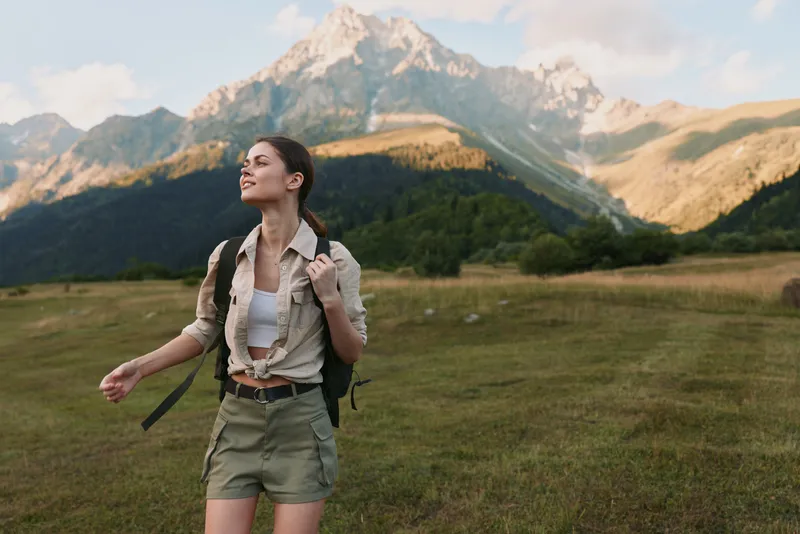 Young woman hiking through a expansive grassy meadow with a backpack, gazing up at distant snow capped mountains in a bright outdoor landscape.