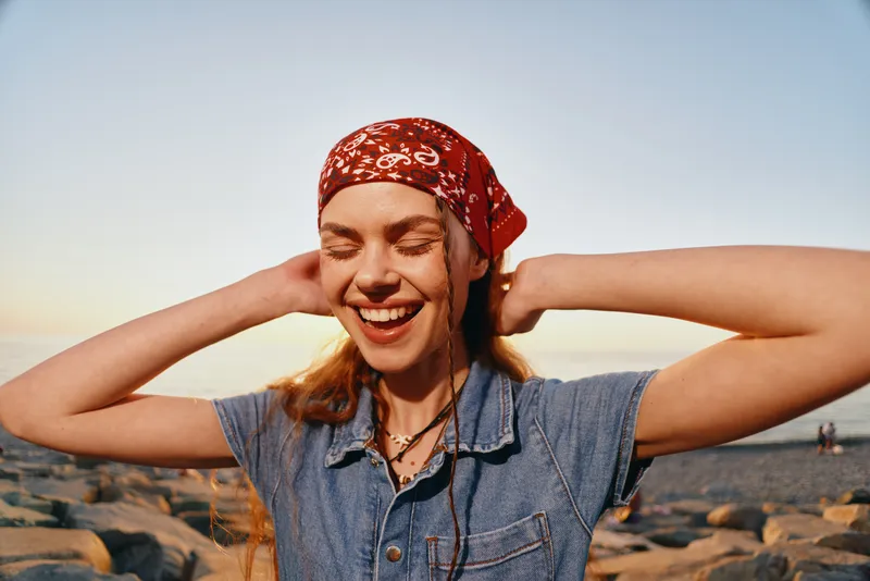 Lifestyle woman in modern boho-western denim street style, smiling with eyes closed outdoors at sunset, wearing a red bandana and casual layered necklaces.