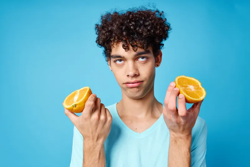 Весёлый парень с кудрявыми волосами держит апельсин на синем фоне