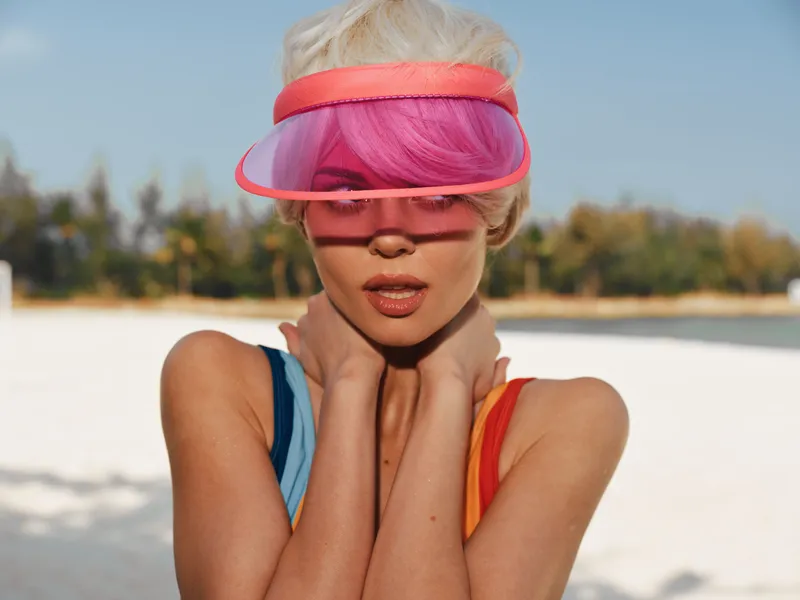 A fashion forward beach portrait featuring a stylish model wearing a pink visor and vibrant hair, sunlit sand and calm sea creating a bold summer vibe.
