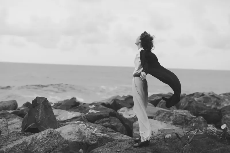 Woman on jagged rocks by the sea, cape billowing in wind, monochrome scene of freedom and resilience. Ideal for travel, fashion, editorial, or lifestyle storytelling with dramatic atmosphere