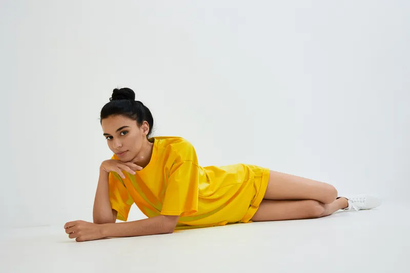 Young woman lying on white floor in bright yellow dress with casual white sneakers, relaxed pose and thoughtful expression in minimal studio setting with soft lighting.