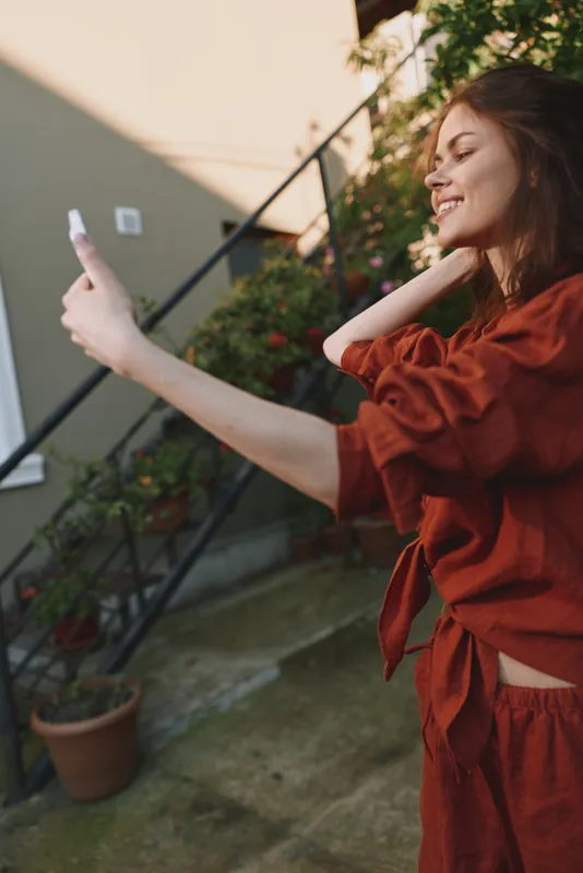Smiling woman in casual orange outfit taking a selfie outdoors. Close-up on a sunlit courtyard with a relaxed lifestyle, candid moment, and contemporary fashion captured in natural light
