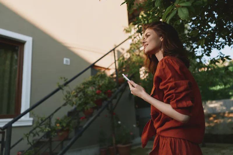 A woman in a red sweater walks outdoors on a set of stairs beside a house, looking at her phone with a relaxed, thoughtful expression, capturing a casual lifestyle moment