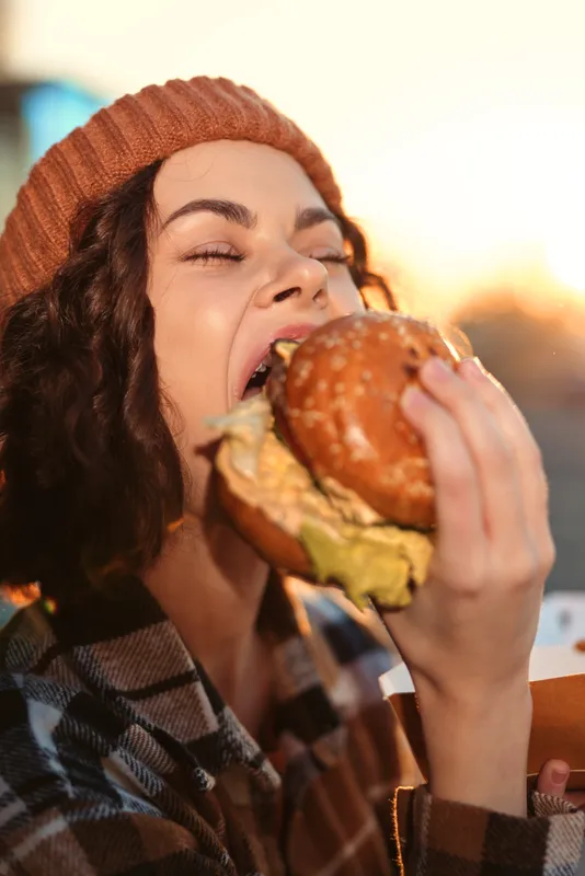 Young woman eating a burger outdoors at sunset, beanie and plaid coat, candid lifestyle shot with golden hour glow, authenticity and emotional storytelling of mindful living and savoring food.
