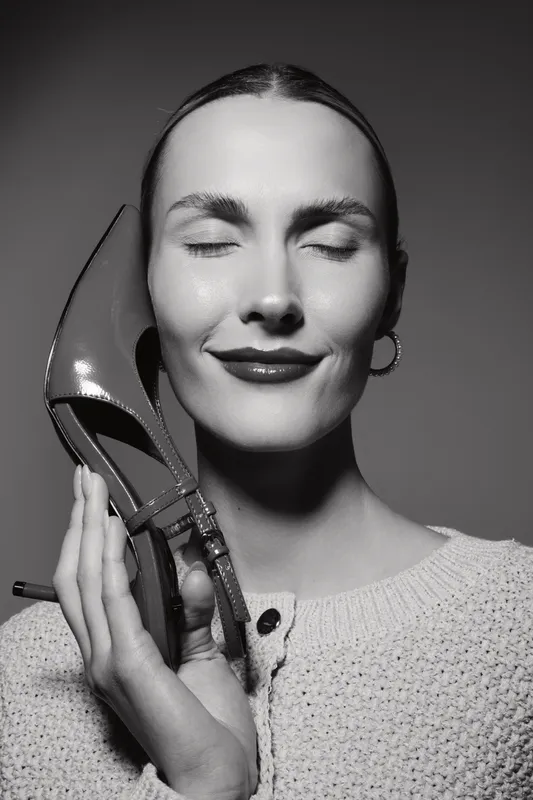 Young white woman with fair skin and dark hair holds high heel shoe near face, eyes closed, smiling with pleasure. Concept for fashion, footwear, beauty, and happiness.