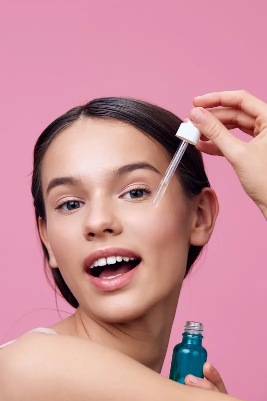 Young woman applying serum for skincare, smiling joyfully on a pink background blending beauty and health, perfect for wellness and skincare concepts