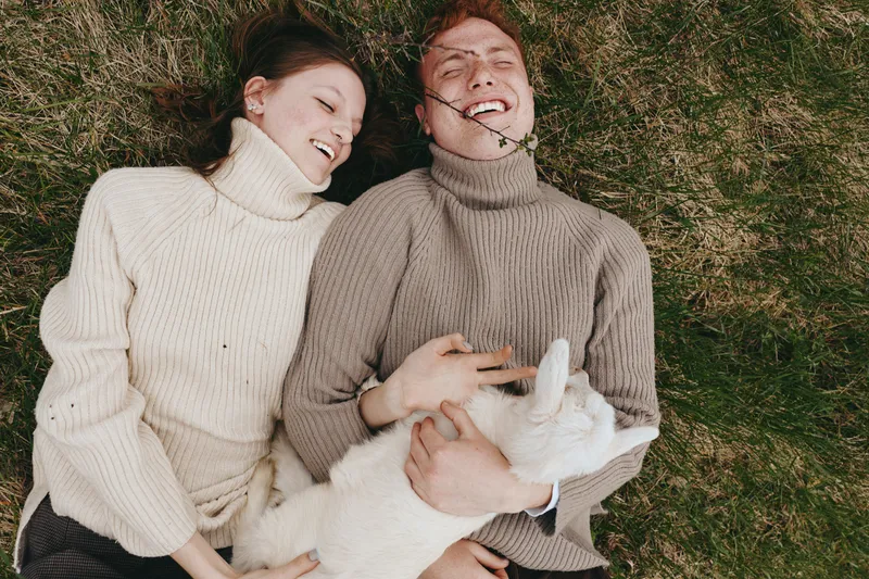 Two smiling adults lie on green grass wearing chunky sweaters, enjoying a cozy outdoor moment together with a small animal between them, showcasing lifestyle comfort, warmth, and carefree affection.
