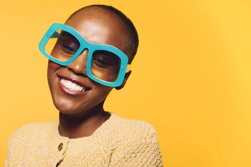 Smiling black woman with dark skin and shaved head wearing large turquoise glasses and beige sweater against yellow background. Concept of happiness, fashion, and confidence in portrait.