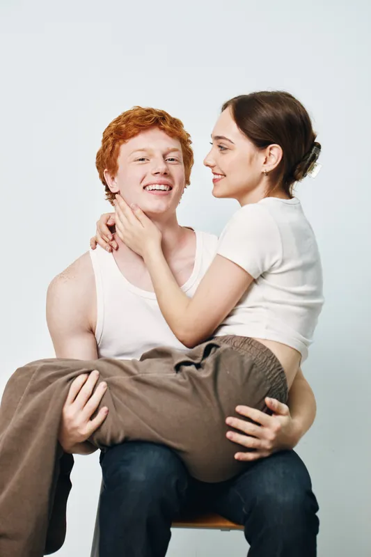 Young couple sitting and smiling with woman in white shirt on man lap in casual clothes with affectionate gesture on gray background studio.