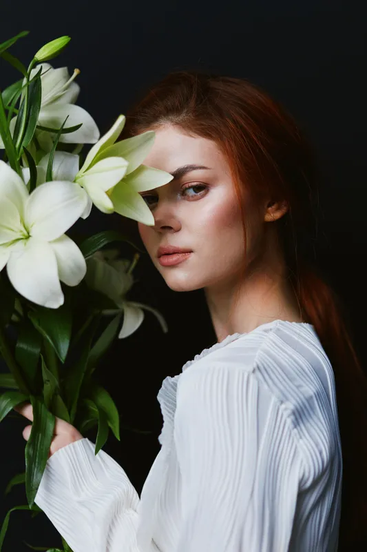 Elegant portrait of a young woman with red hair holding white lilies, wearing a light blouse, against a dark background, conveying beauty and serenity