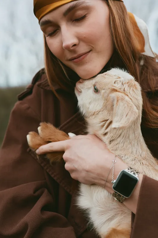 Warm moment with a woman embracing a goat kid outdoors, soft light, calm expression, cozy attire, natural surroundings, affection, nature, lifestyle, relaxed posture, candid outdoors