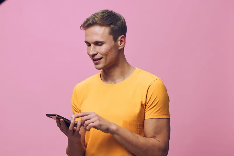 Smiling young man wearing bright yellow t-shirt using smartphone isolated on pink background. Casual people lifestyle concept, cheerful expression, technology, communication, modern life.