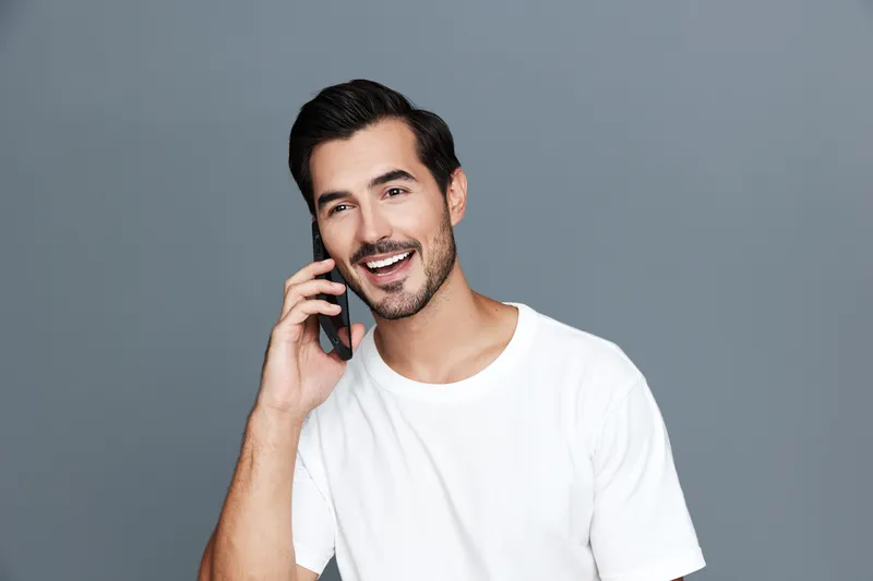 Happy smiling young man talking on mobile phone, wearing a casual white t-shirt, standing against a plain gray background. Lifestyle and communication concept.