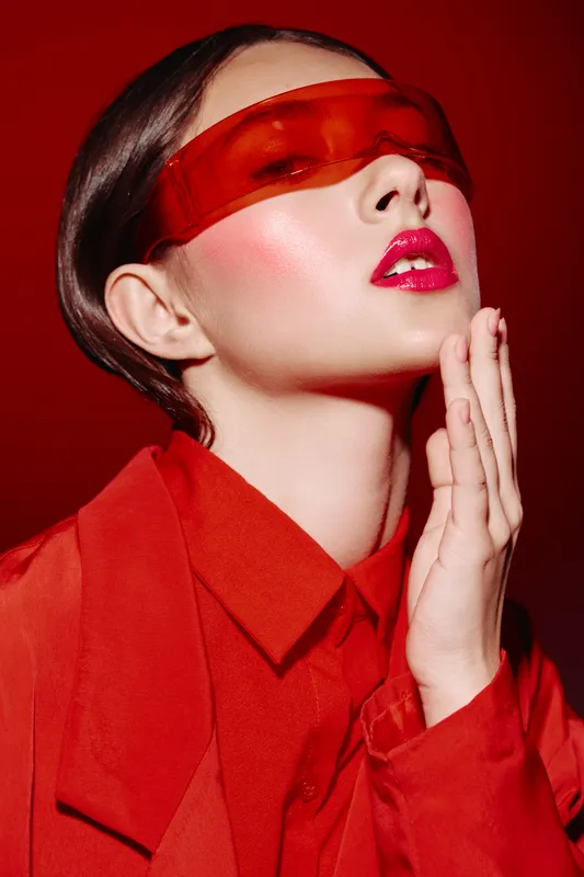 Young woman in bright red fashion jacket and matching red visor with glossy lipstick. She gently touches her chin inside studio with red background. Beauty concept for trend and style.