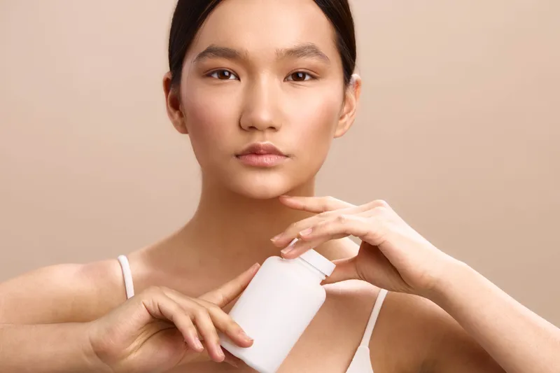 Asian woman applies skincare product in a clean, minimal setting, her calm expression and smooth complexion conveying care, wellness, and a mindful beauty routine.