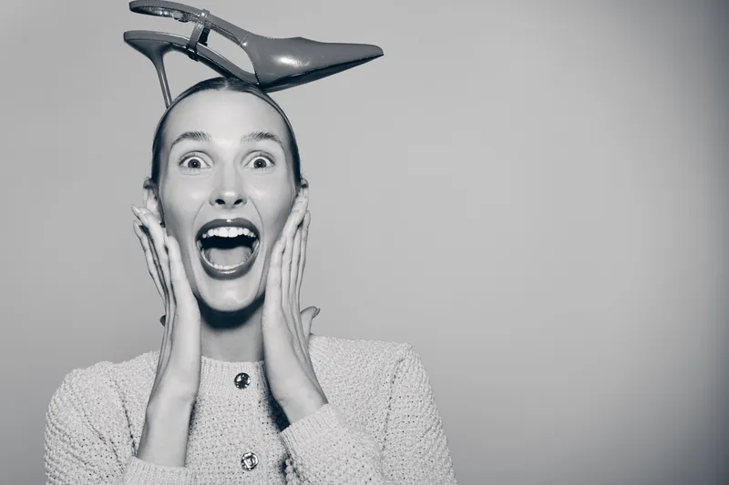 Excited white woman with pale skin holds high heel shoe on head, smiling widely. Studio portrait with playful mood and fashion concept on grey background.
