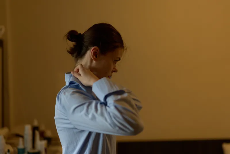 A young woman in a blue shirt stands indoors rubbing her neck with a pained expression, suggesting discomfort or stress in a softly lit room.