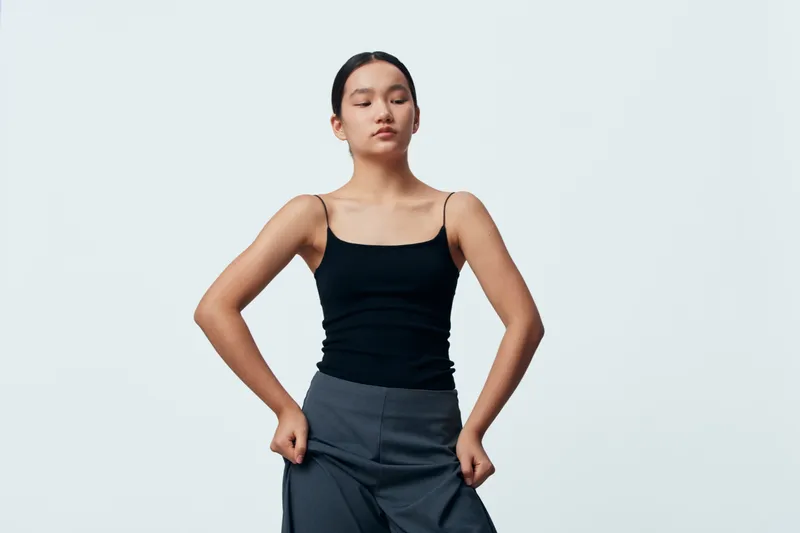 Young woman in black tank top and gray pants standing with eyes closed and hands in pockets against plain background. Casual fashion portrait with calm expression and relaxed pose.