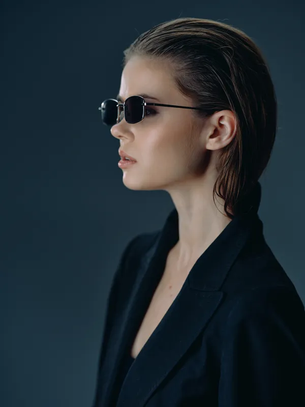 Stylish woman wearing sunglasses and black blazer posing on dark background with serious expression