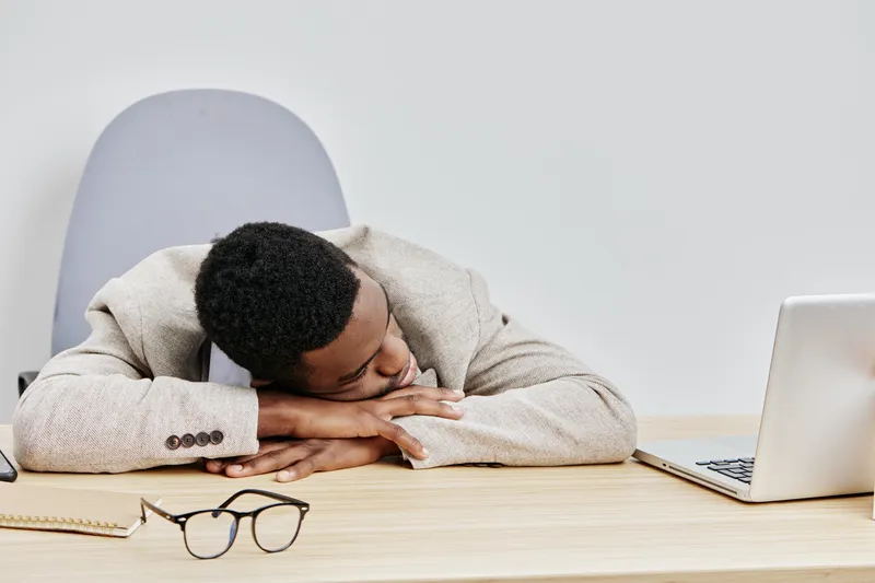 Relaxed young African-American man resting his head on his desk while working on a laptop in a modern office setting, with glasses and notebook nearby. People lifestyle concept, office work, tiredness