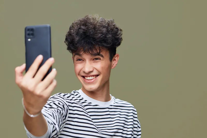 smiling young man takes a selfie with smartphone, curly hair, striped shirt, joyful expression, light green background, casual style portrait.