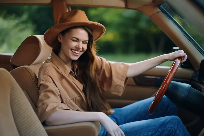 Happy young woman in a brown shirt and hat, smiling joyfully while driving a classic car on a sunny day in nature, symbolizing freedom and adventure.