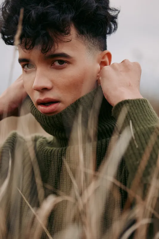Close-up portrait of a young man with short curls wearing a green knit turtleneck, conveying authenticity and credibility through a sincere, grounded expression.