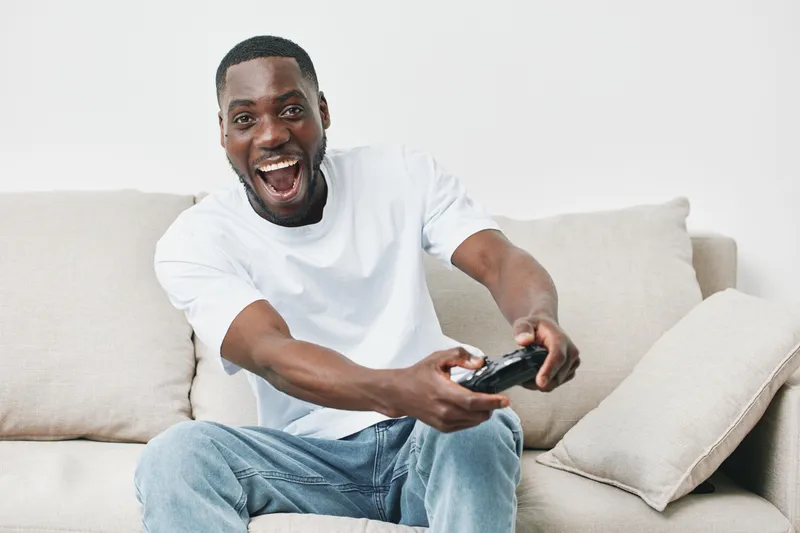 Excited African American man joyfully playing video games on a beige sofa, showcasing a vibrant and lively home atmosphere