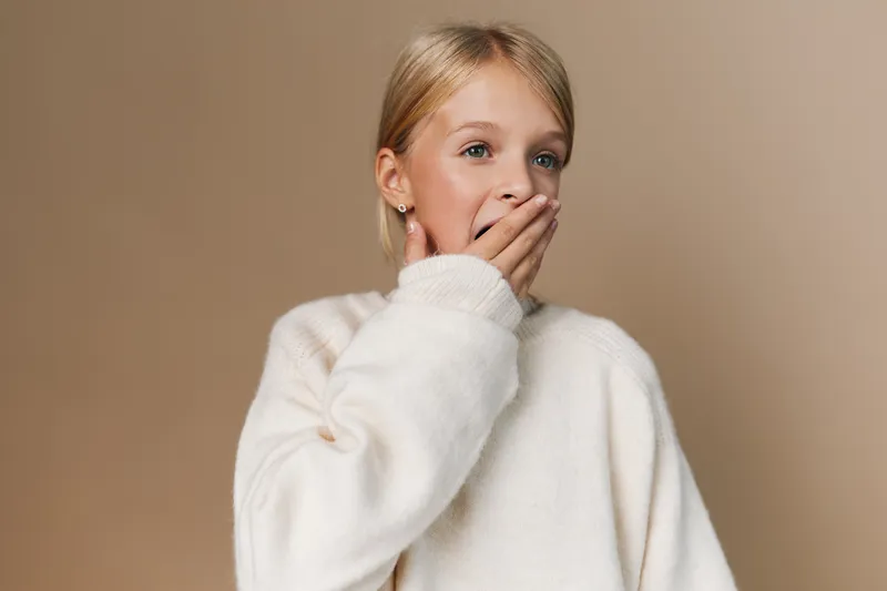 Youthful woman with blonde hair wearing a cozy white sweater, covering her mouth in a surprised gesture, against a warm beige backdrop with soft lighting and texture.