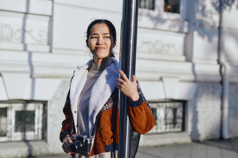 Woman leaning on a lamppost outdoors with a camera, wearing a scarf and jacket, natural light, candid street moment, casual style and relaxed mood