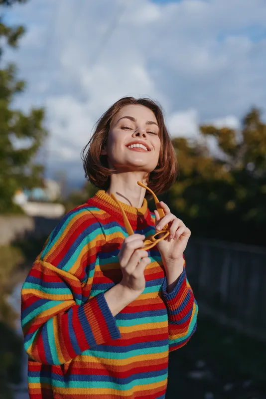 Woman smiling in a colorful striped sweater and sunglasses, casual outfit, short hair outdoor portrait in golden hour light, joyful expression and confident mood for lifestyle and fashion.